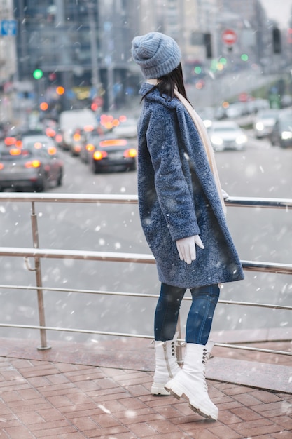 Mulher morena atraente usa um casaco de inverno da moda e um chapéu, olhando para o trânsito da cidade à noite durante a nevasca.