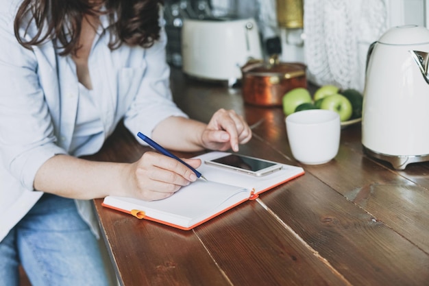 Mulher morena casual fazendo anotações no livro diário com e usando celular na cozinha em casa