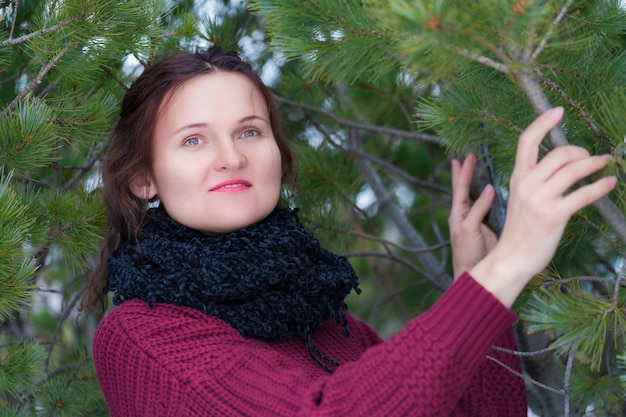 Mulher morena com cabelo comprido e olhos castanhos, vestida com um pulôver marrom e lenço preto no pescoço, em pé em uma floresta de pinheiros