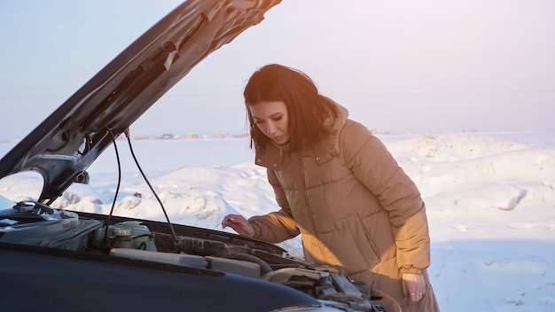 Mulher morena com casaco quente olha para o motor de um automóvel quebrado com o capô aberto em pé na estrada de neve na zona rural em um dia ensolarado de inverno