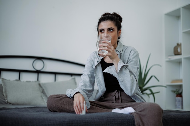 Mulher morena com distúrbio mental a beber água e a segurar comprimidos no quarto de casa.