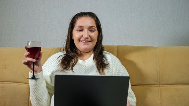 Mulher morena com suéter de malha branca toma pequenos goles de vinho e fala com entusiasmo por meio de videochamada usando laptop, ficando em casa isolado.