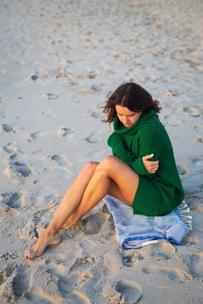 Mulher morena com um suéter verde senta-se perto da praia e admira o mar.