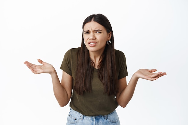 Foto mulher morena confusa olhando perplexa para a frente, encolhendo os ombros e estendendo as mãos para os lados, sem noção, não sei o que a pessoa quer, parede branca