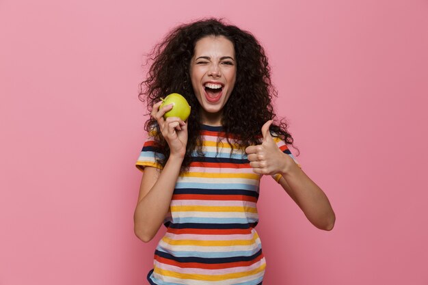 Mulher morena de 20 anos com cabelo encaracolado sorrindo e segurando uma maçã verde isolada na rosa