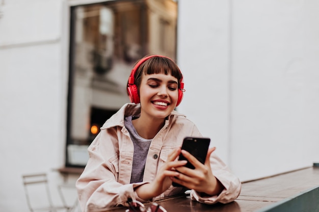 Mulher morena de jaqueta bege segurando o telefone e ouvindo música lá fora