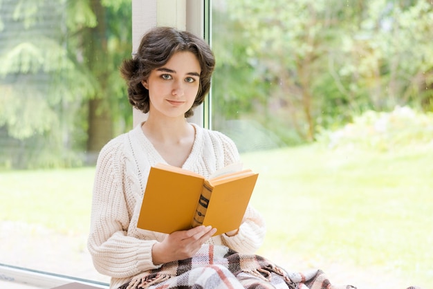 Mulher morena de suéter quente lendo livro sentado no quarto na varanda