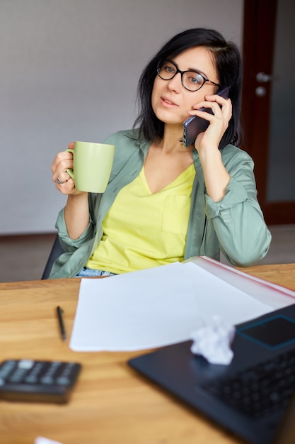 Foto mulher morena elegante de óculos, sentada à mesa de madeira com o bloco de notas e recebendo uma ligação, bebe chá no moderno local de trabalho