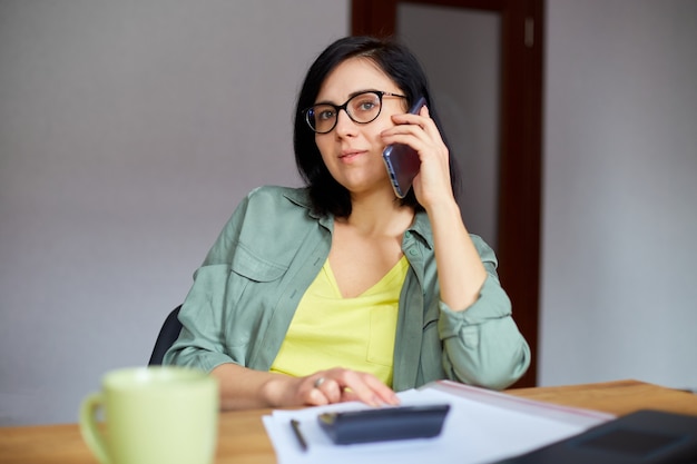 Mulher morena elegante de óculos, sentada à mesa de madeira com o bloco de notas e recebendo uma ligação no ambiente de trabalho moderno.