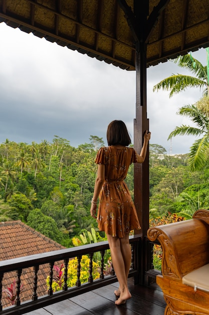 Mulher morena encantada em pé perto do suporte enquanto pensa e observa a paisagem tropical