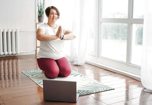 Mulher morena encantadora adulta plus size corpo positivo prática ioga com laptop em casa