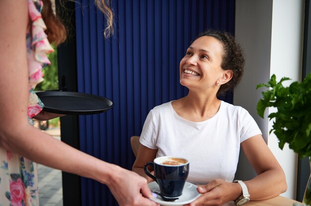 Mulher morena encantadora sentada à mesa no café e sorrindo para a garçonete servindo e trazendo uma xícara com cappuccino.