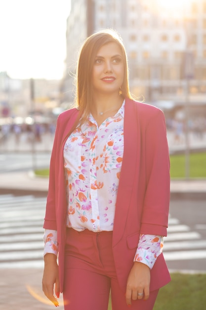 Mulher morena encantadora vestindo blusa e jaqueta rosa, posando para o fundo da cidade à noite.