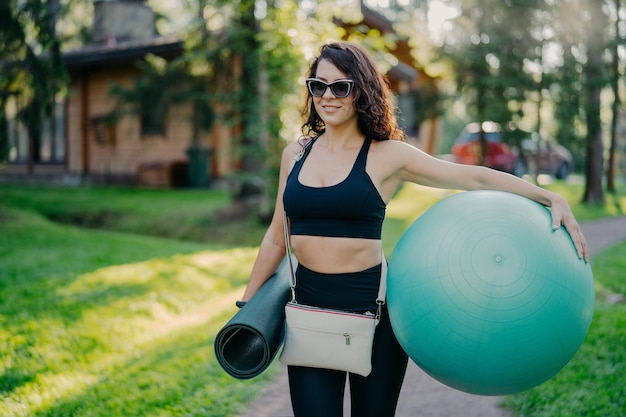 Mulher morena encaracolada feliz esportiva usa top cortado e perneiras segura bola de fitness e karemat enrolado vai ter exercícios de fitness em poses ao ar livre contra casa particular e grama verde