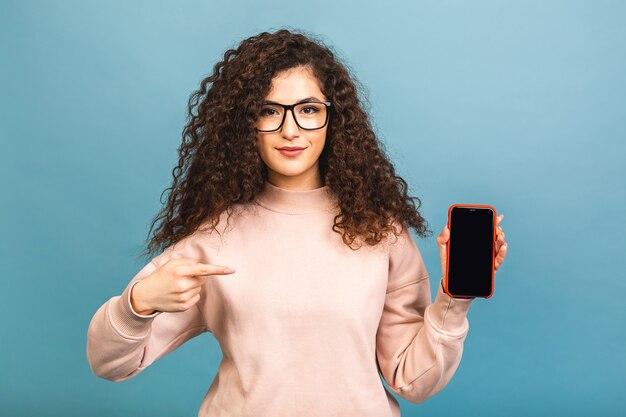 Mulher morena encaracolada feliz surpresa mostrando a tela do smartphone em branco e apontando para ela enquanto olha para a câmera