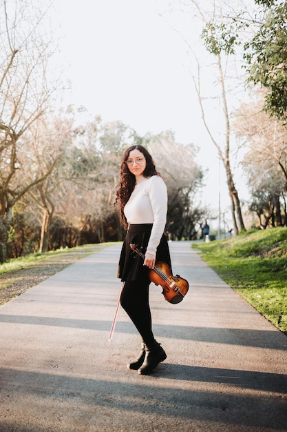 Mulher morena encaracolada segurando um violino e um arco, em pé na estrada do parque ao pôr do sol.