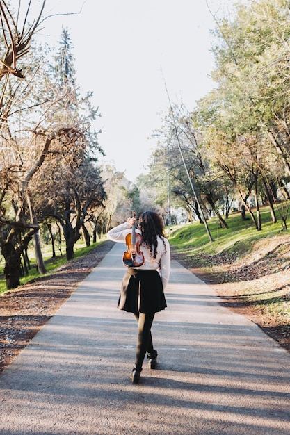 Foto mulher morena encaracolada segurando um violino nas costas caminhando pela estrada do parque ao pôr do sol