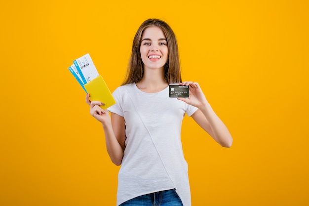 Mulher morena feliz com bilhetes de avião no passaporte e cartão de crédito isolado sobre amarelo