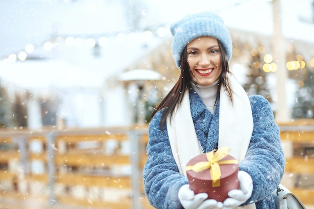 Mulher morena feliz com casaco de inverno, segurando uma caixa de presente na feira de natal, durante a queda de neve. espaço para texto