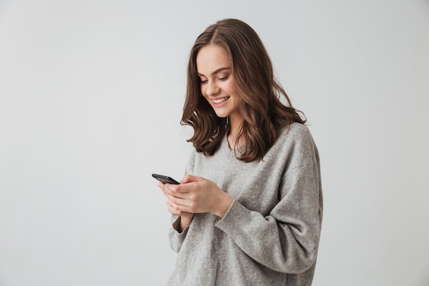 Mulher morena feliz com suéter escrevendo mensagem no smartphone sobre uma parede cinza