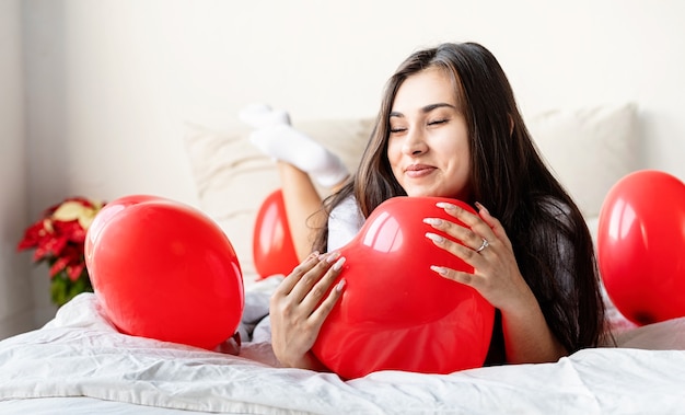 Mulher morena feliz deitada na cama com balões em forma de coração vermelho