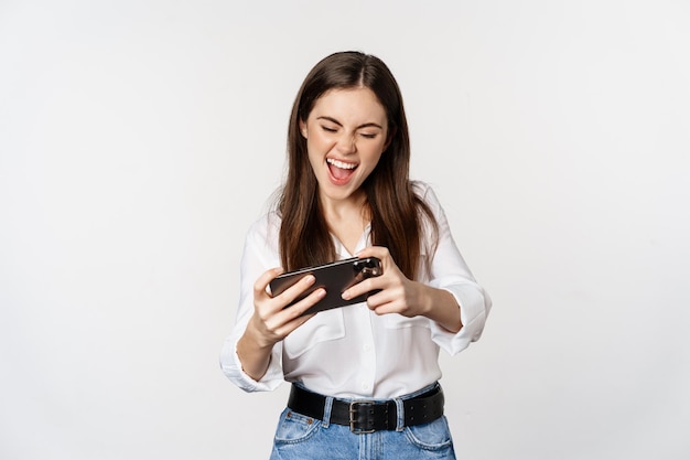 Mulher morena feliz jogando videogame para celular sorrindo e olhando para a tela animada em pé sobre...