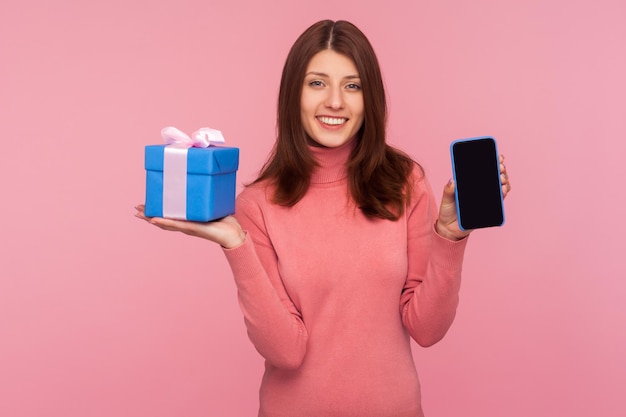 Mulher morena feliz no suéter rosa segurando e mostrando o telefone celular e a caixa de presente azul Foto de estúdio interior isolada em fundo rosa