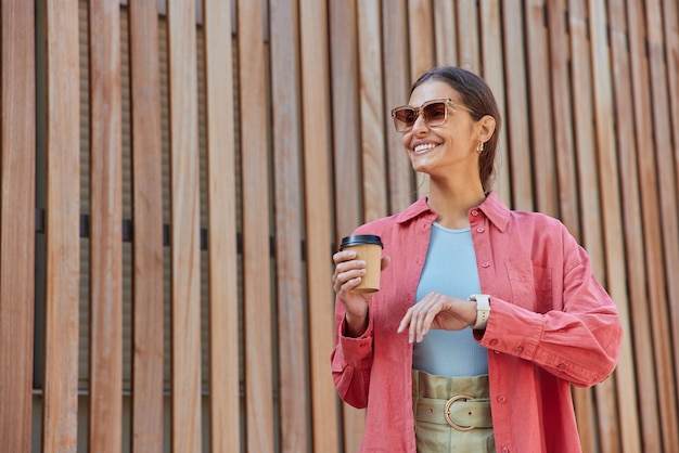 Mulher morena feliz positiva sorri e desvia o olhar bebe café para viagem gosta de bom dia usa óculos de sol camisa rosa e relógio de pulso posa contra madeira, portanto, simula espaço para sua promoção