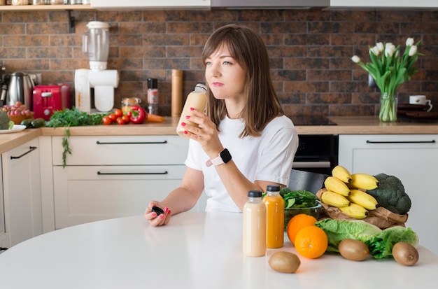 Mulher morena feliz sentada com garrafa de smoothie e frutas na cozinha de casa Refeição vegana e conceito de desintoxicação Garota com camiseta branca bebendo coquetel fresco Maquete de embalagem