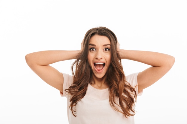 Foto mulher morena feliz surpresa em t-shirt gritando e cobrindo os ouvidos enquanto sobre parede branca