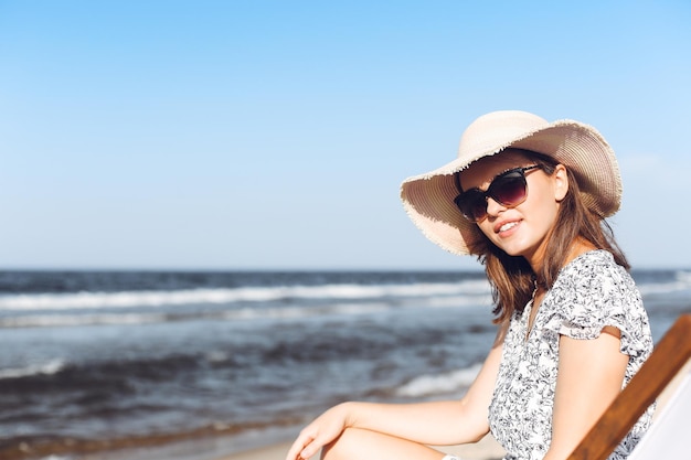 Mulher morena feliz usando óculos escuros e chapéu relaxando em uma cadeira de deck de madeira na praia do oceano