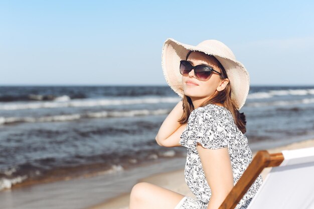 Mulher morena feliz usando óculos escuros e chapéu relaxando em uma cadeira de deck de madeira na praia do oceano.