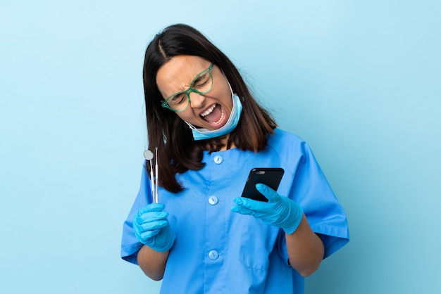 Foto mulher morena jovem dentista de raça mista segurando ferramentas sobre fundo isolado com telefone em posição de vitória
