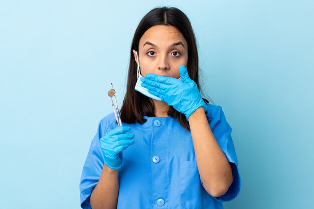 Mulher morena jovem dentista de raça mista segurando ferramentas sobre isolado parede cobrindo a boca com as mãos