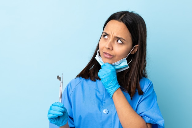 Mulher morena jovem dentista de raça mista segurando ferramentas sobre isolado tendo dúvidas