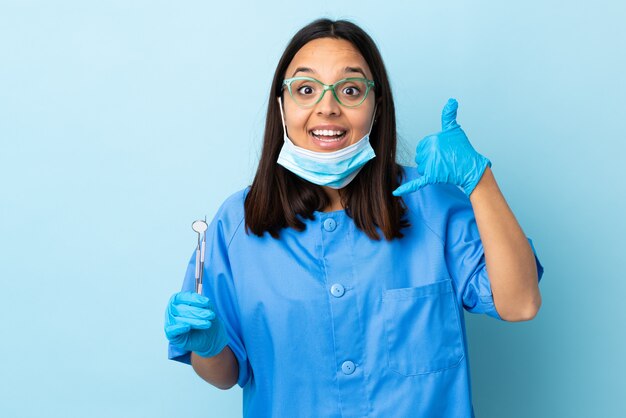 Mulher morena jovem dentista de raça mista segurando ferramentas sobre parede isolada, fazendo gesto de telefone e duvidar