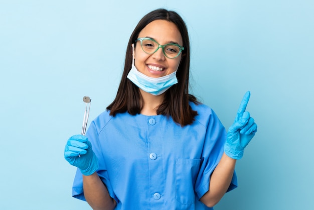 Mulher morena jovem dentista de raça mista segurando ferramentas sobre parede mostrando e levantando um dedo em sinal dos melhores