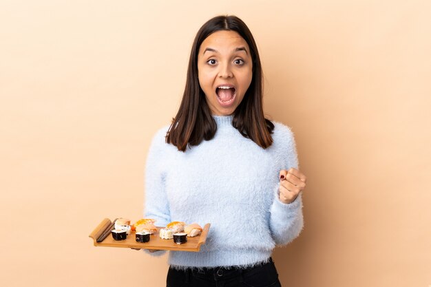 Foto mulher morena jovem raça mista segurando sushi sobre fundo isolado, comemorando uma vitória na posição de vencedor