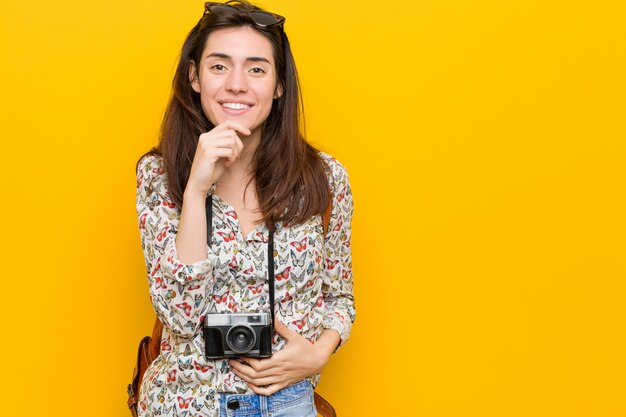 Mulher morena jovem viajante sorrindo feliz e confiante, tocando o queixo com a mão.