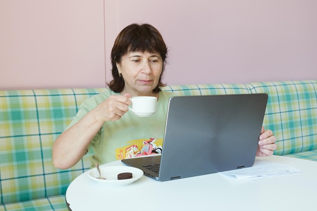 Mulher morena madura trabalha na frente de um laptop e bebe café