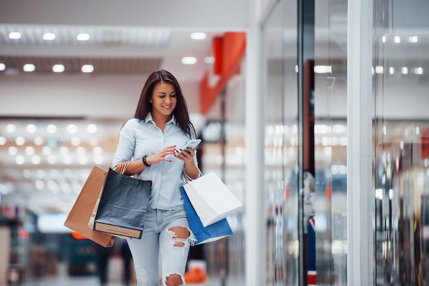 Mulher morena no supermercado com muitos pacotes e telefone nas mãos tem dia de compras.