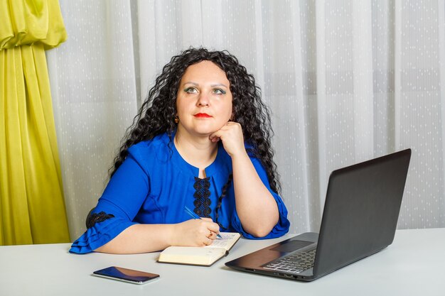 Mulher morena pensativa e encaracolada está sentada à mesa no escritório. Foto horizontal