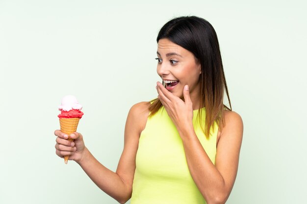 Mulher morena segurando um sorvete de corneta sobre parede verde com surpresa e expressão facial chocada
