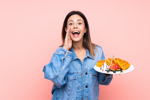 Mulher morena segurando waffles sobre parede rosa gritando com a boca aberta