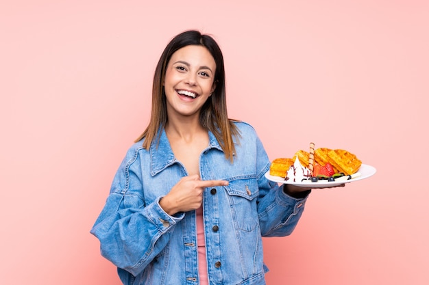 Mulher morena segurando waffles sobre parede rosa isolada e apontando-o