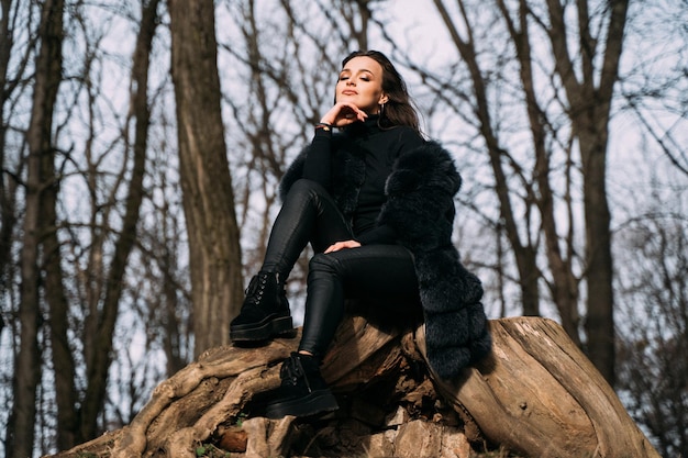 Mulher morena sentada em uma pedra no parque Modelo feminino sexy em calças pretas elegantes e casaco de pele pensando e sonhando