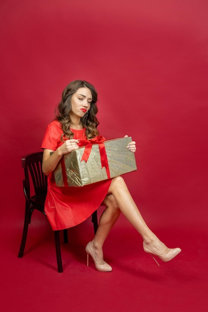 Mulher morena sentada na cadeira desapontada com o presente em vestido vermelho sobre fundo vermelho