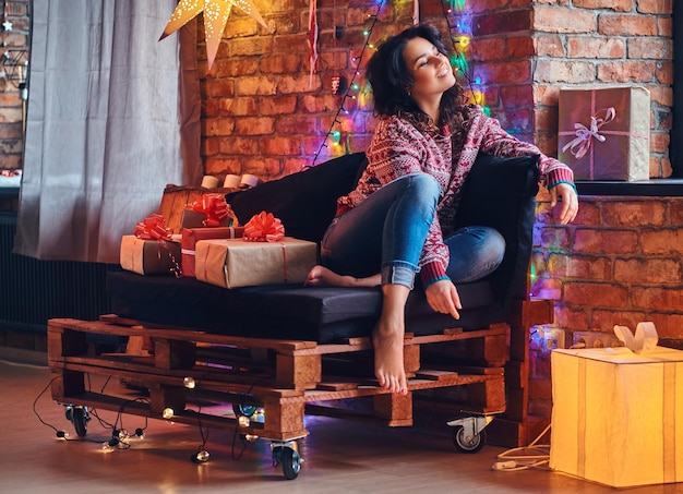 Mulher morena sexy com pé nu, vestida de jeans e um suéter vermelho, posando em um sofá em uma sala com decoração de natal.