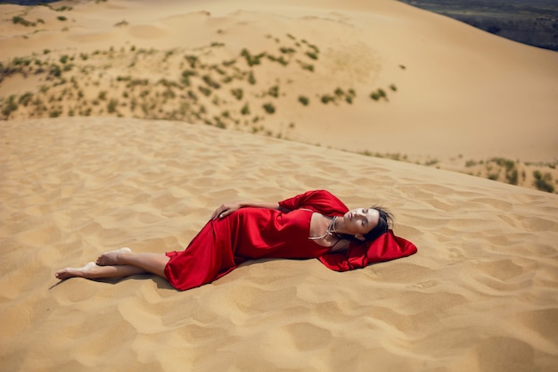 Mulher morena sexy com um vestido longo vermelho sentada em uma duna no verão no Daguestão