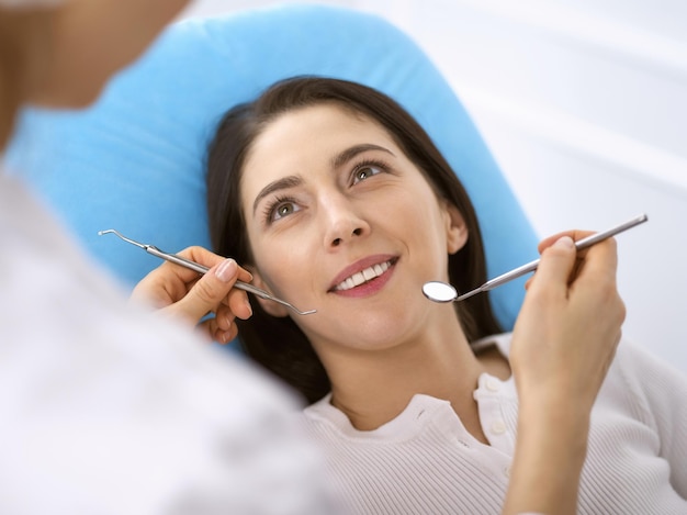 Mulher morena sorridente sendo examinada pelo dentista na clínica odontológica. Mãos de um médico segurando instrumentos odontológicos perto da boca do paciente. Dentes saudáveis e conceito de medicina.